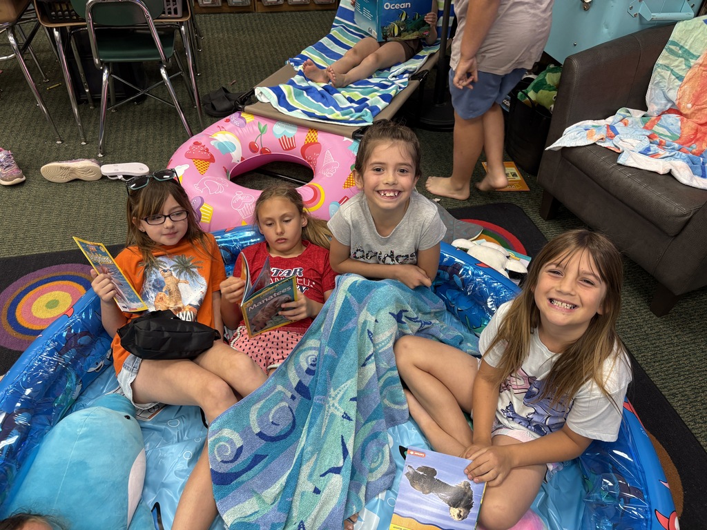 Four children, seated in an inflatable pool, hold books and appear to be reading. The pool is blue.