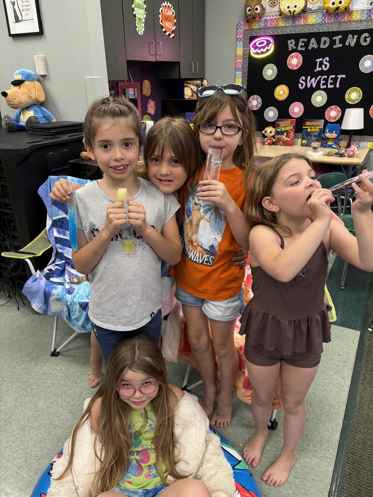 Five young girls are standing and sitting in a room with a chair and decorations. They hold snacks and drinks.