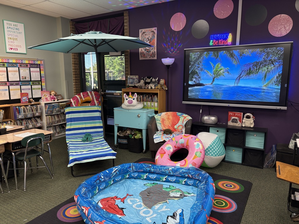 Interior of a colorfully decorated classroom with purple walls and round decorations. A TV displays a beach scene, and a beach-themed inflatable pool sits on the floor.