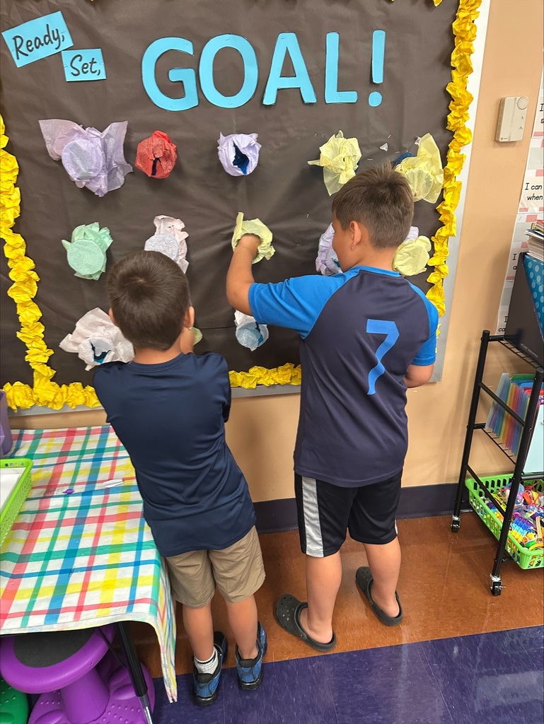 Two young boys in front of a bulletin board with "GOAL!" written in blue. One boy is reaching for a paper.