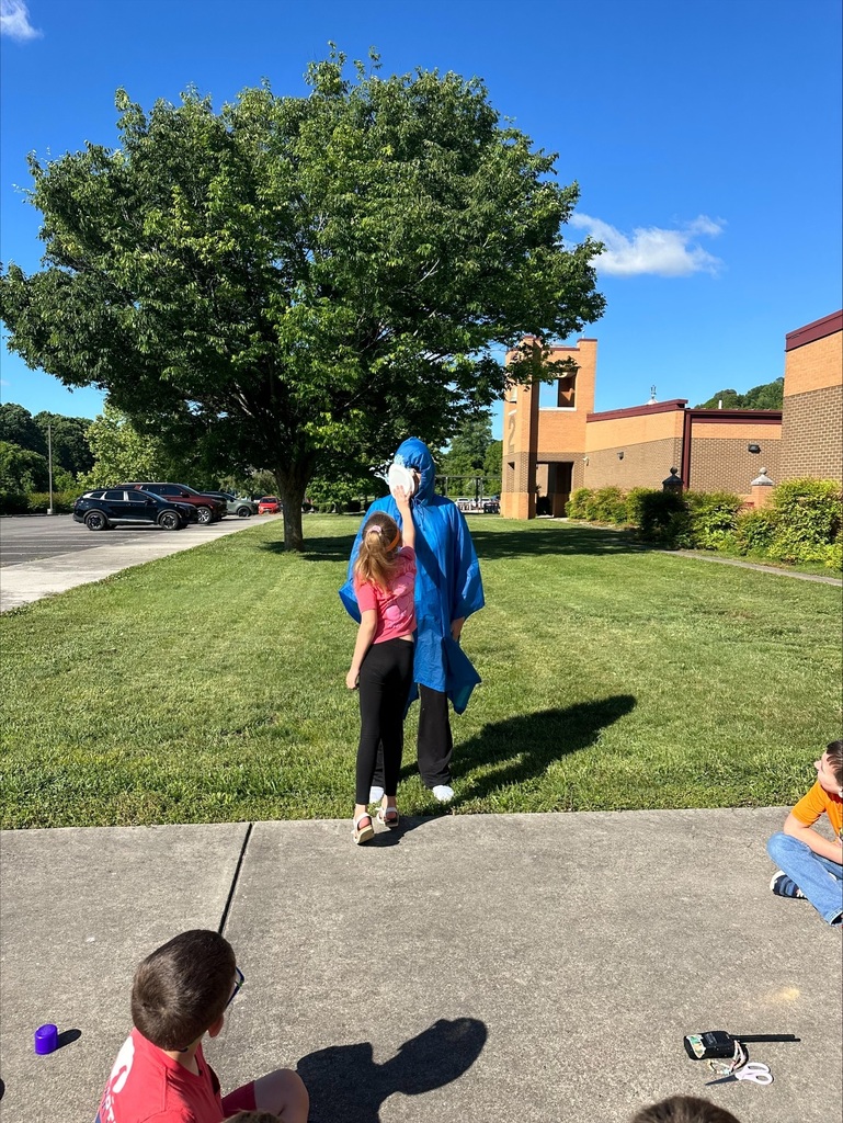 Two people, one in a blue cape, walk on grass; a child sits on a sidewalk.
