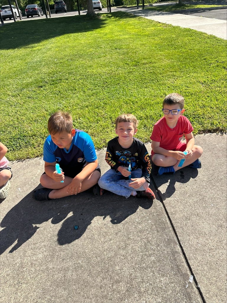 Three boys sit on a sidewalk, holding drinks. One wears glasses. A grassy area and parked cars are in the background.