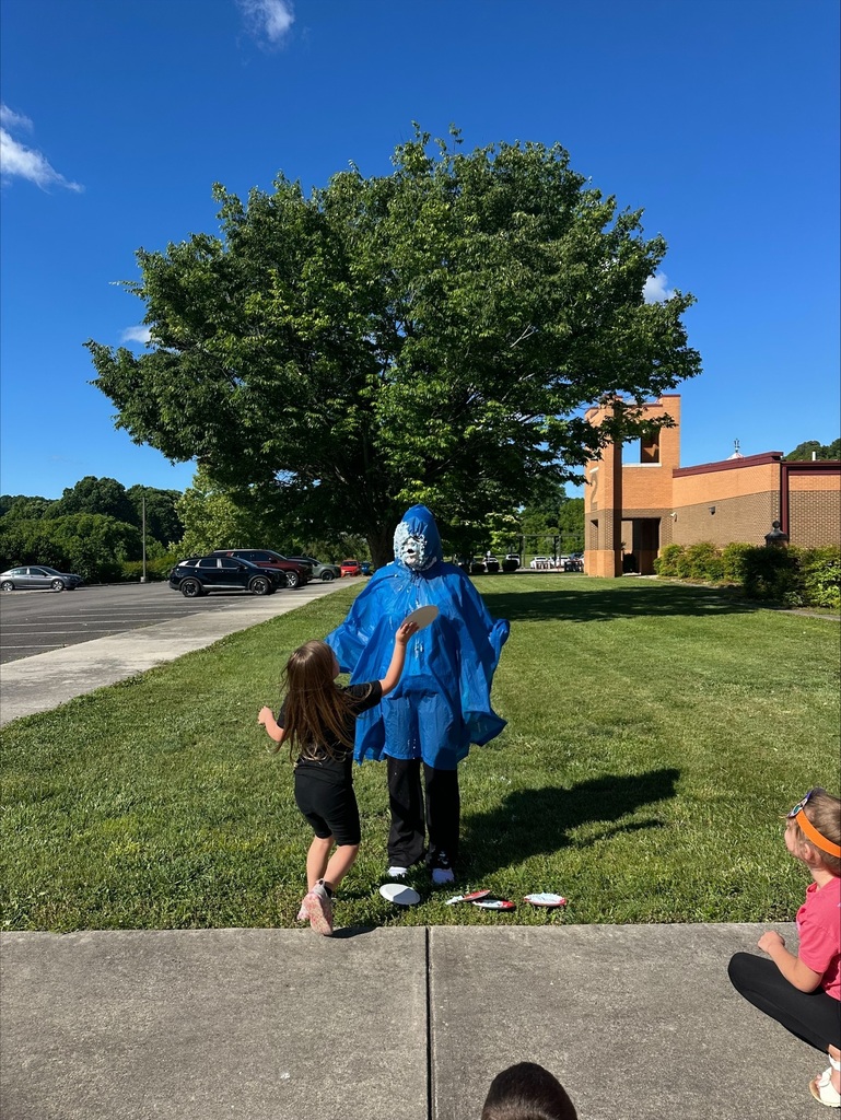 A person in a blue cape stands near a young child. A child sits on the sidewalk. Trees and a building are in the background.