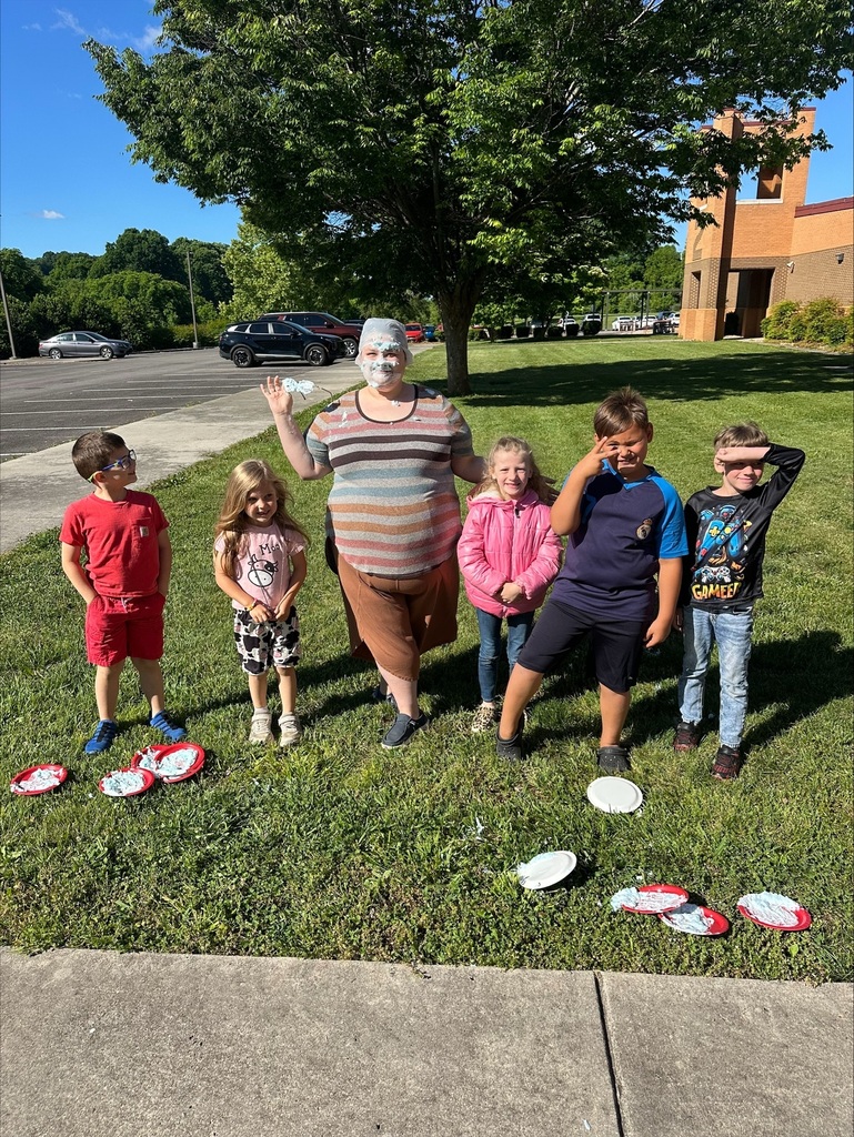 A group of children and an adult stands on grass with plates and a tree in background.