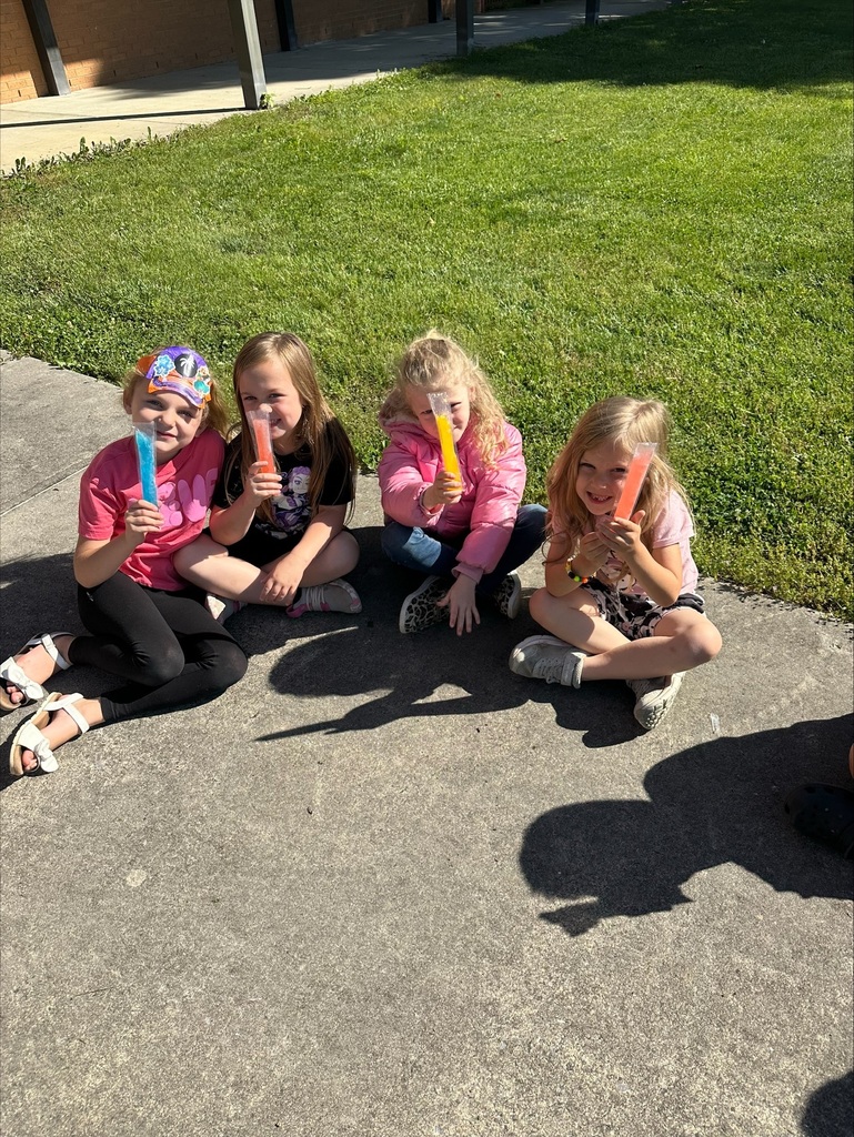 Four young girls are sitting on the sidewalk. They are holding popsicles in various colors. Green grass is behind them.
