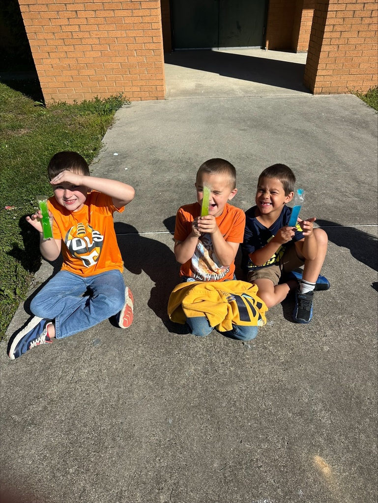 Three boys in orange shirts sit on a sidewalk, holding blue objects and smiling. Behind them is a brick building.