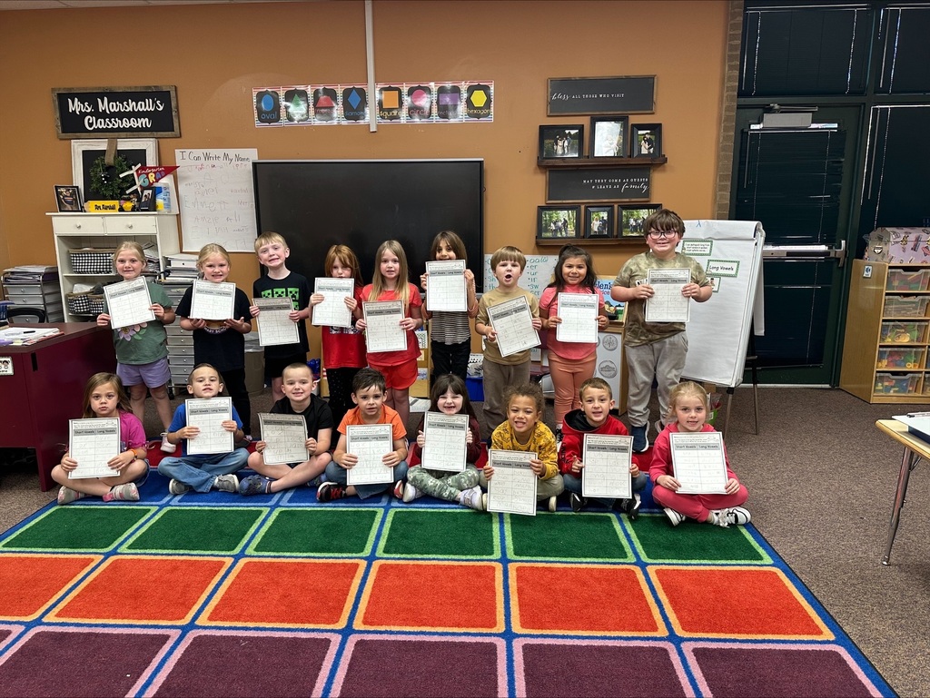 Group of young children in a classroom holding certificates, seated on a colorful floor mat.