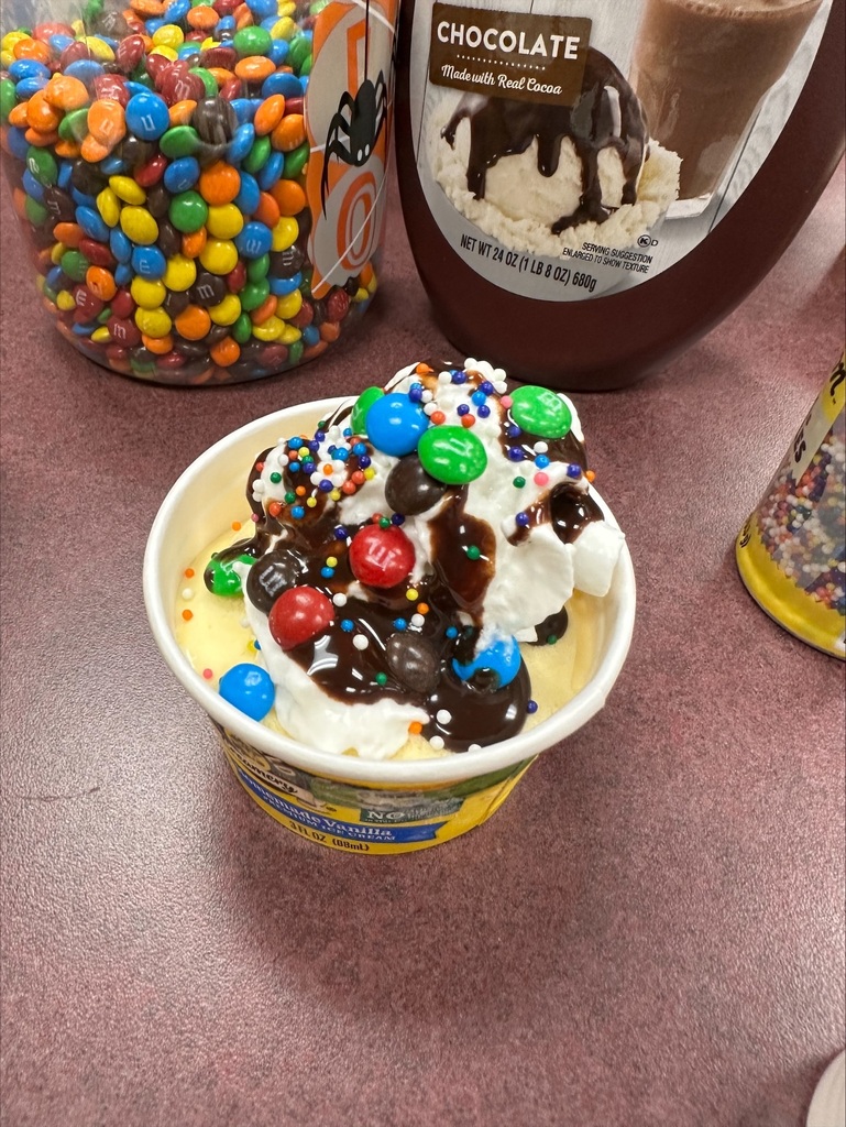 A scoop of ice cream with toppings in a yellow cup. Containers of chocolate syrup and colorful candies nearby.