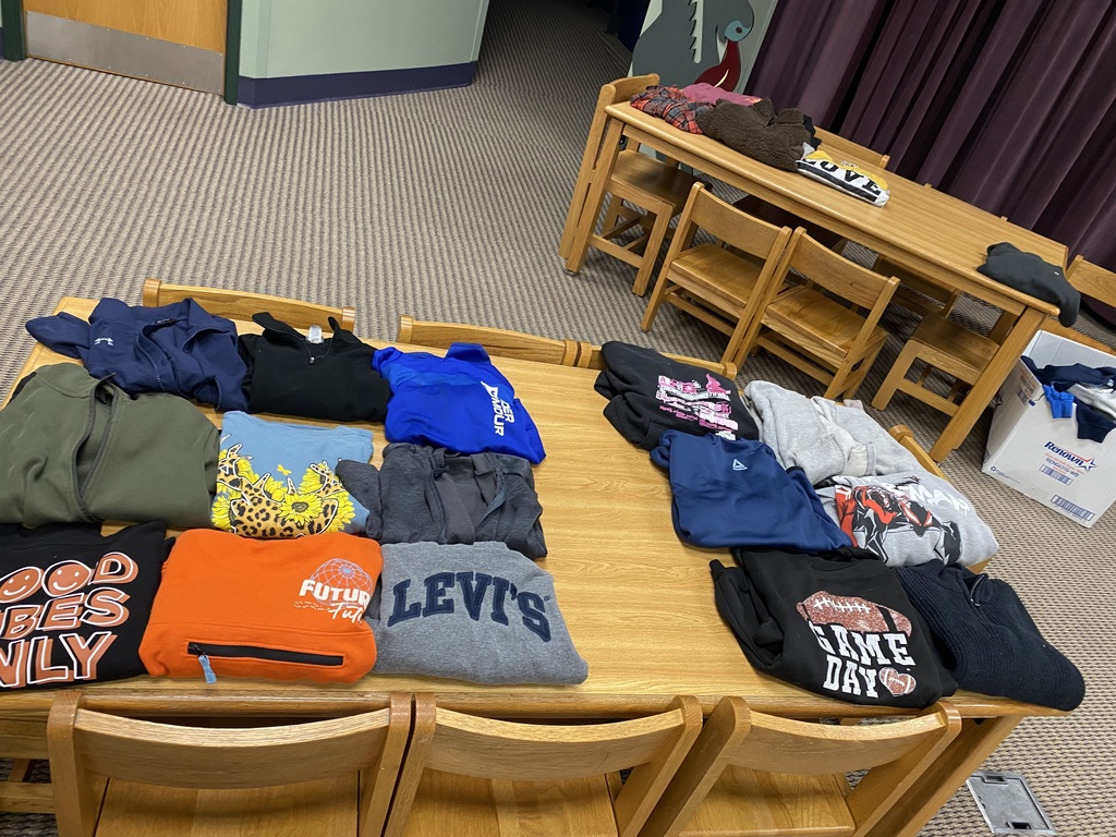 A wooden table with various clothing items, including shirts and hoodies, displayed. A wooden desk with chairs is behind it.