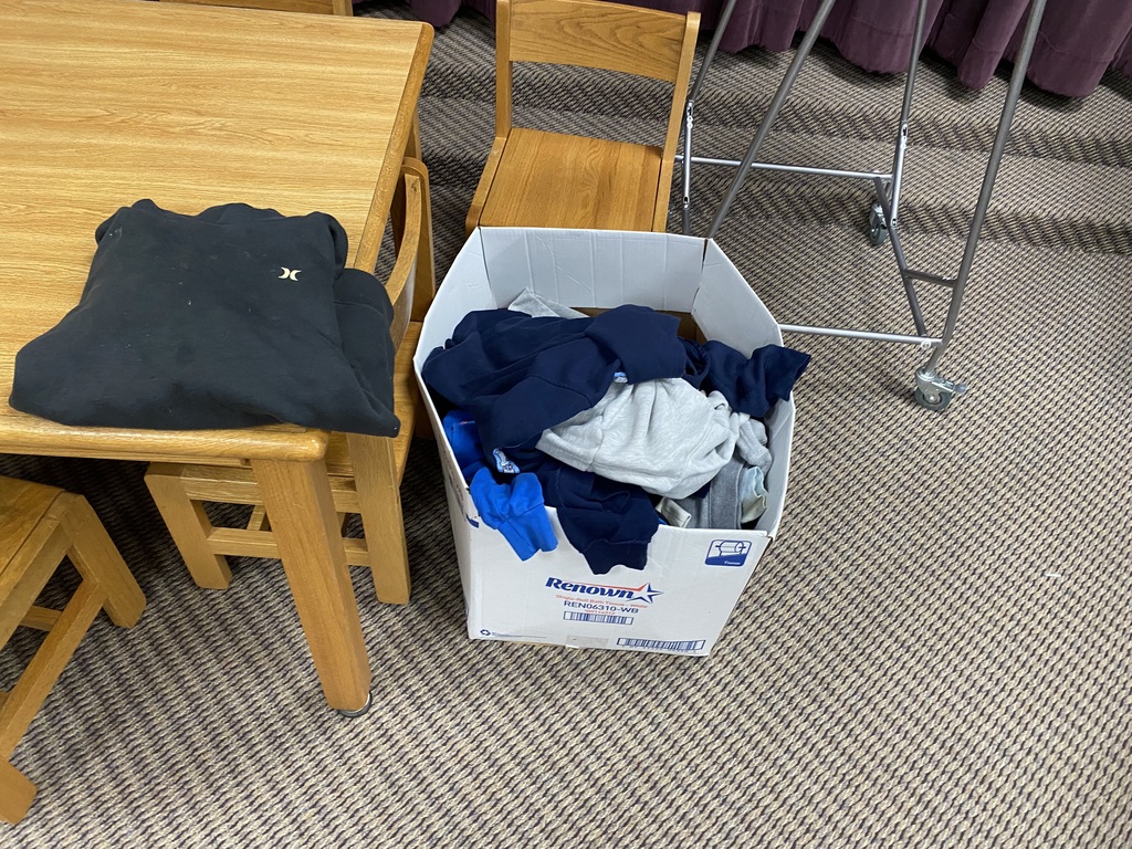 A wooden table has a black t-shirt on it. A white storage box with assorted colored clothes is next to it. A metal clothing rack and wooden chairs are nearby.