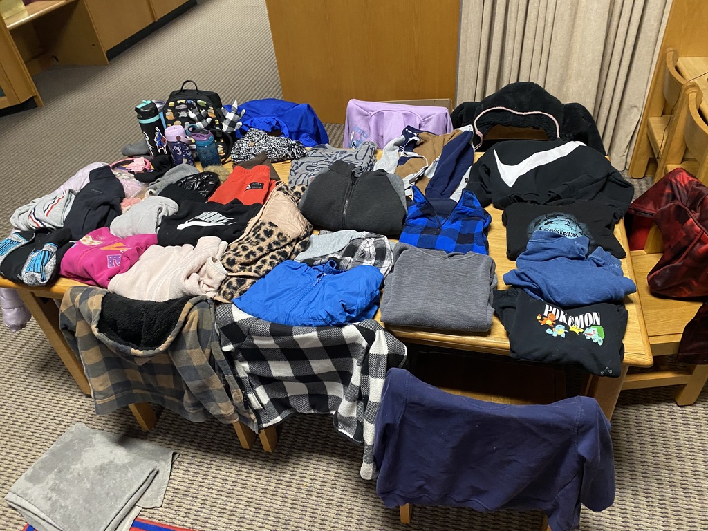 A table with various clothing items on it, including hoodies, jackets, shirts, and hats. The table is surrounded by chairs.