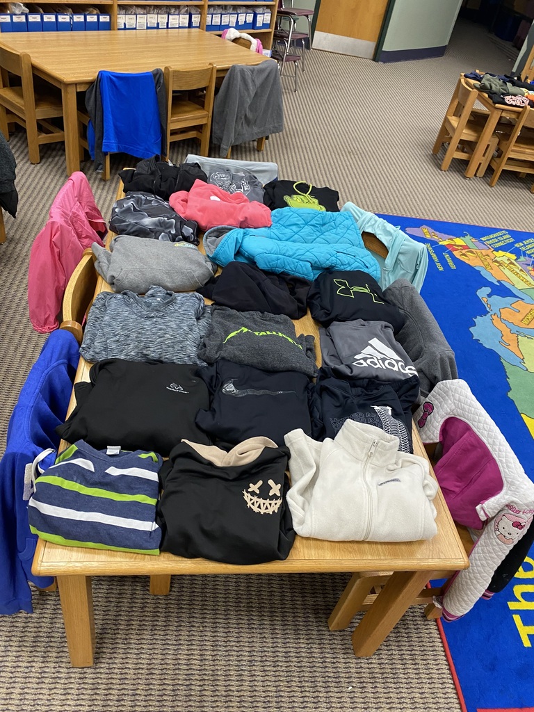 A table with a variety of folded clothing items on it, set in a room with a rug and wooden chairs.