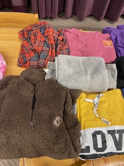 A brown jacket, yellow shirt, pink, purple, and gray t-shirts, and gray hoodies on a wooden table.