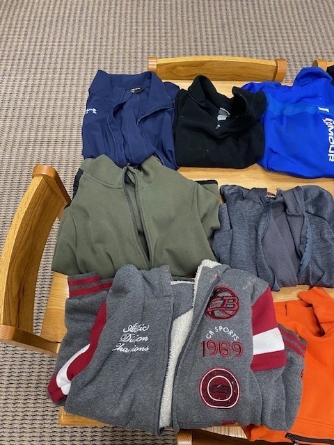 A wooden chair holds assorted clothing on a carpeted floor, including hoodies and T-shirts in various colors and sizes.