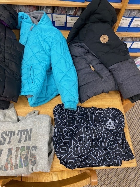 Three jackets and two sweatshirts are displayed on a wooden table. Background includes shelves with storage boxes.
