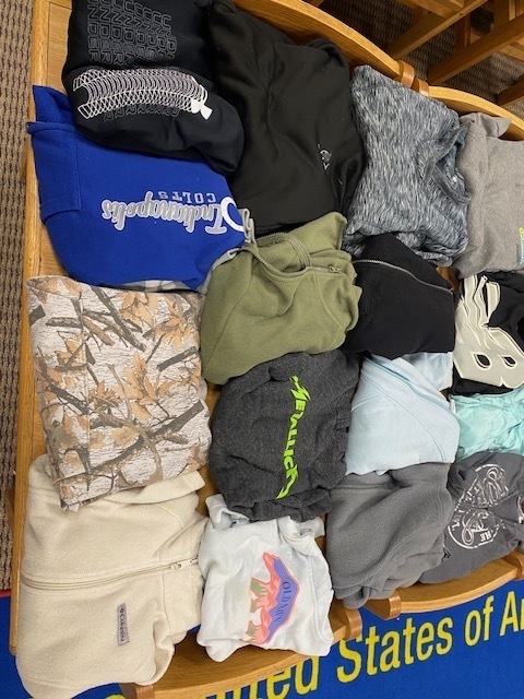 A collection of folded clothing, mostly T-shirts, in various colors, displayed on a wooden shelf.