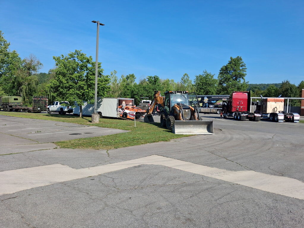 A paved area with a snowplow on grass. Multiple vehicles, including a red truck, are parked. Trees are in the background.