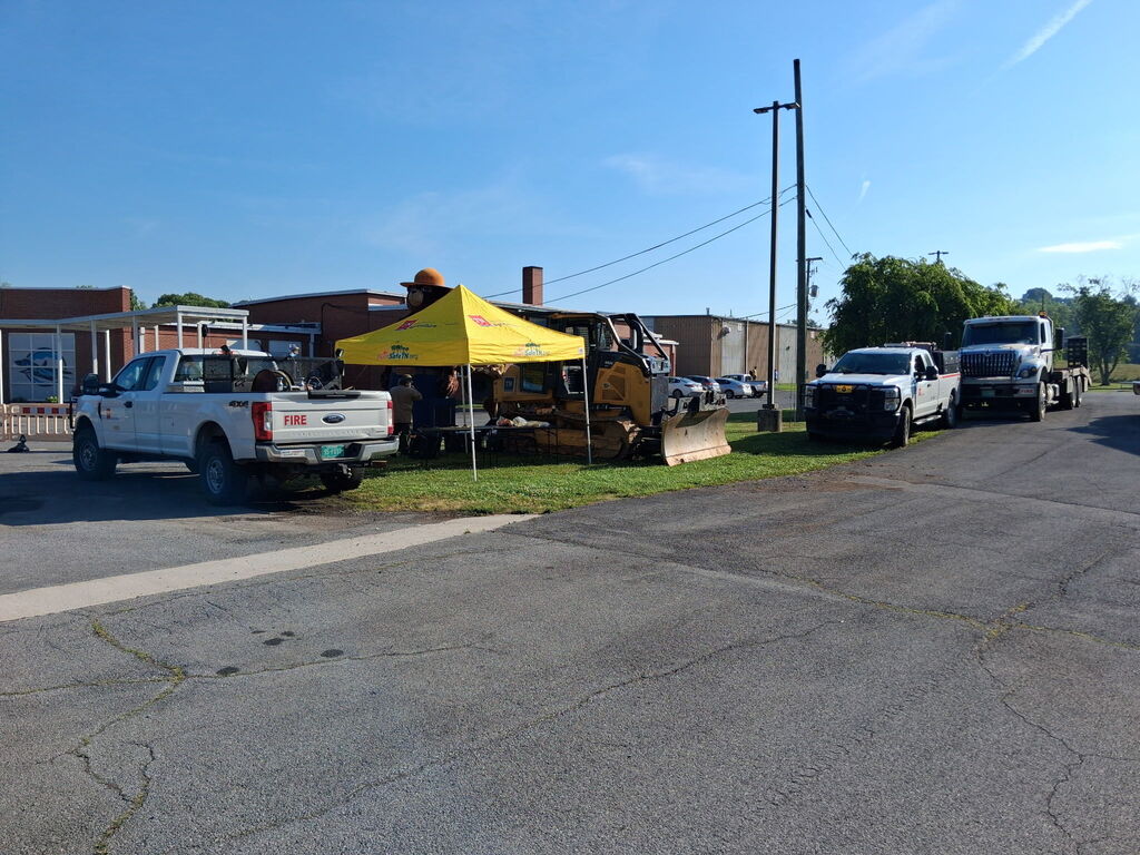 Vehicles, including trucks and a yellow tent, parked by a road. Behind, a building with a chimney.