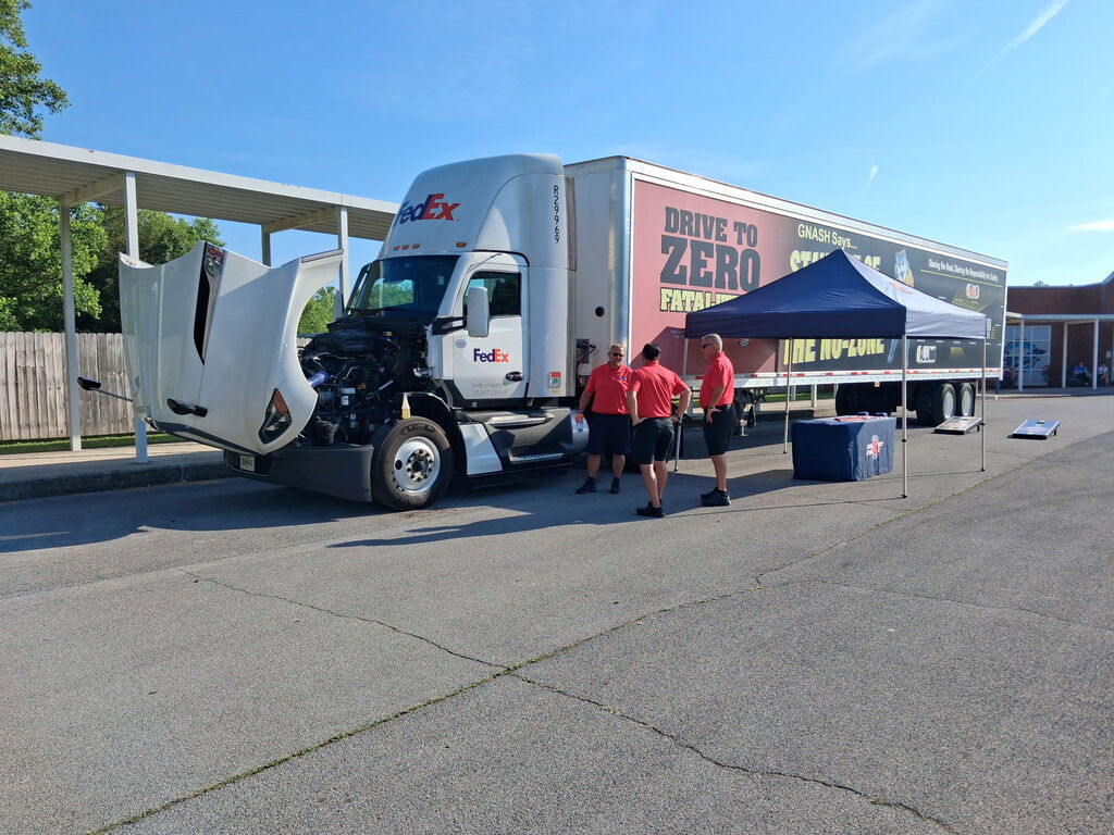 A large white truck with "Drive to Zero" written on its side is parked. Three people stand nearby, two in red shirts.