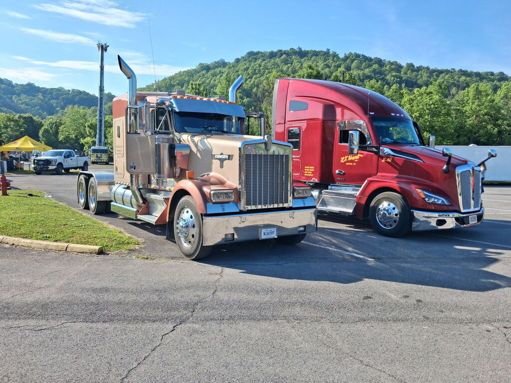 Two large trucks, one brown and one red, parked next to each other in a lot. Background includes trees and mountains.