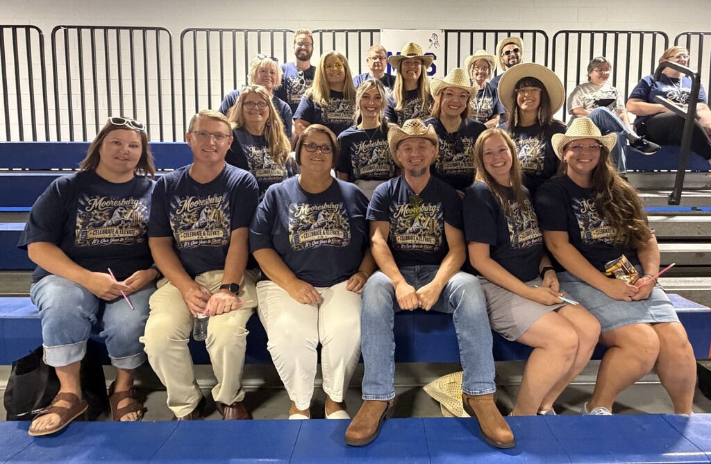 A group of adults wearing blue shirts, hats, and some sunglasses sit together on bleachers.