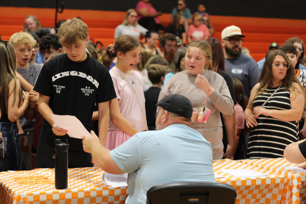 A man reads from a paper at a table, while others stand and watch. Behind, a crowd in a gym.