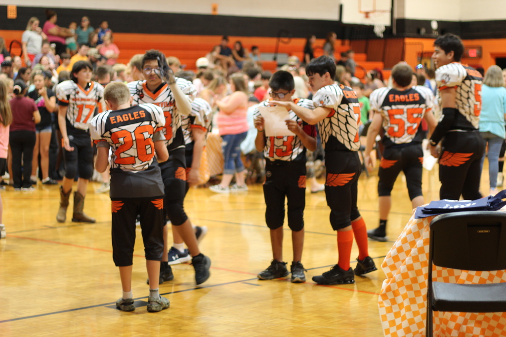 A group of football players wearing jerseys with numbers 26, 55, and 38. They stand in a basketball court with spectators.