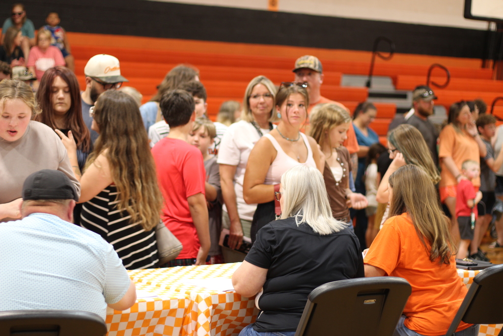 People in a gymnasium, some standing and others sitting at a table with a checkered cover.