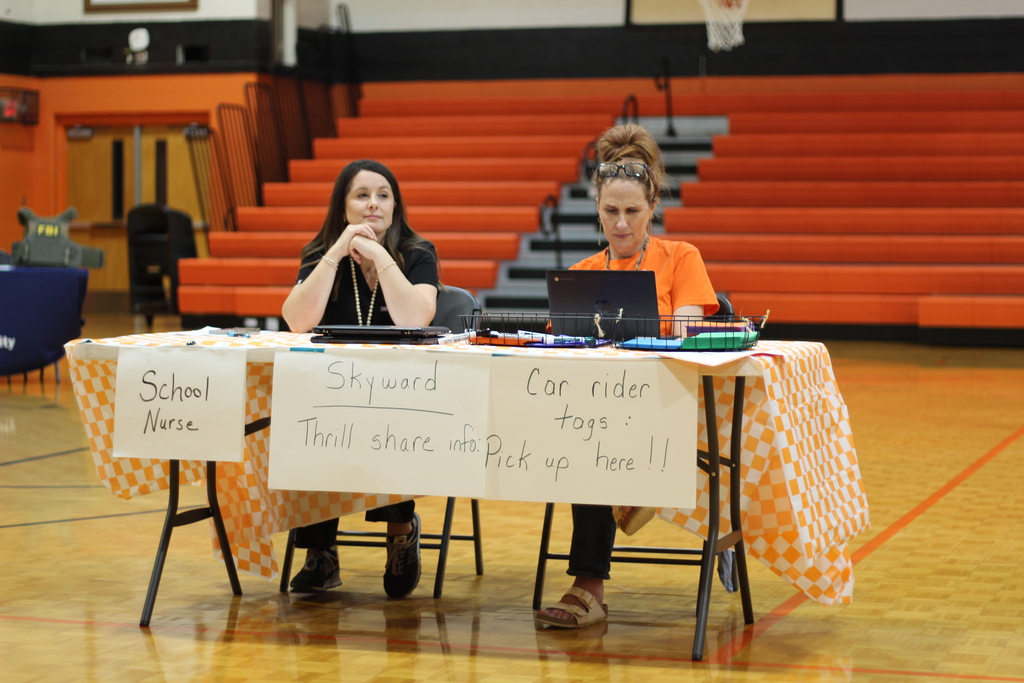 Two women seated at a table in a gym, using laptops. Tablecloth has signs reading "School Nurse" and "Skyward" and "Car rider."