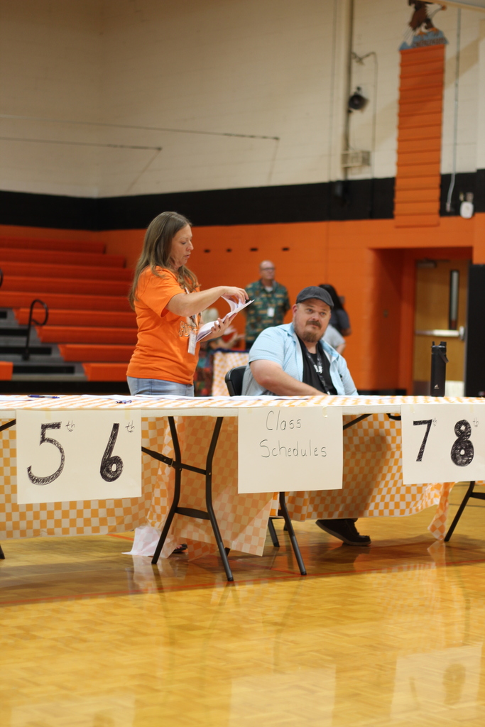 An indoor court with a woman at a table reading from papers. Behind her, a man sits. Tables have signs: "56" and "78".