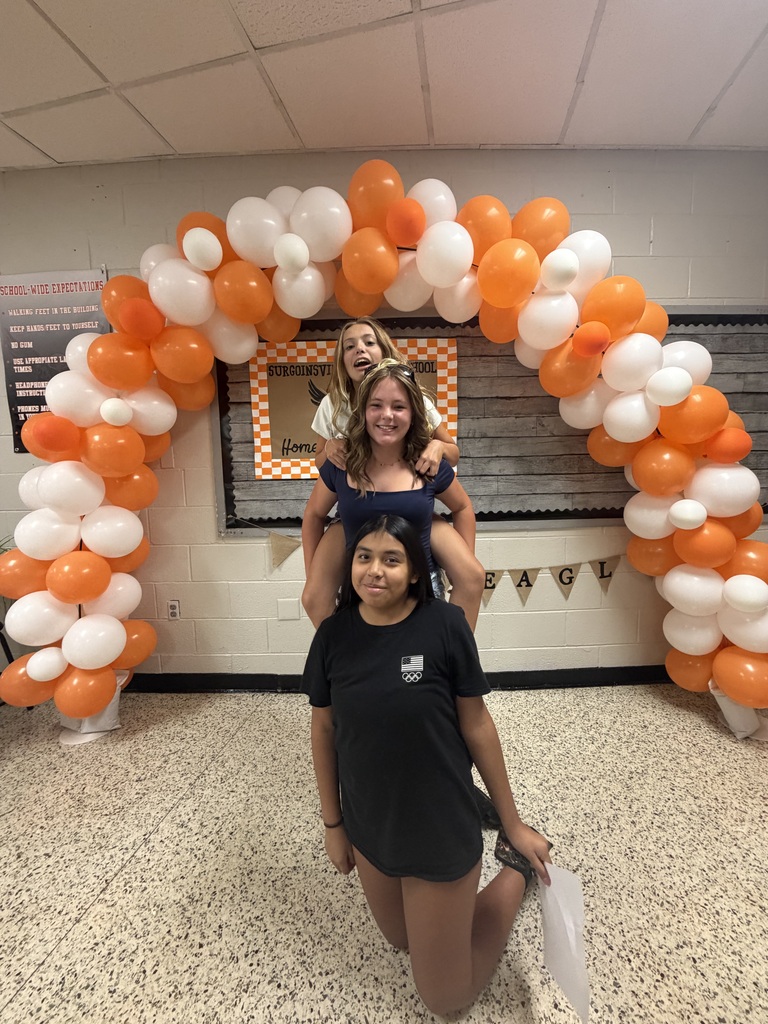 Three individuals are under a balloon arch of orange and white balloons. One is kneeling, one is sitting on another's shoulders, and one is standing.