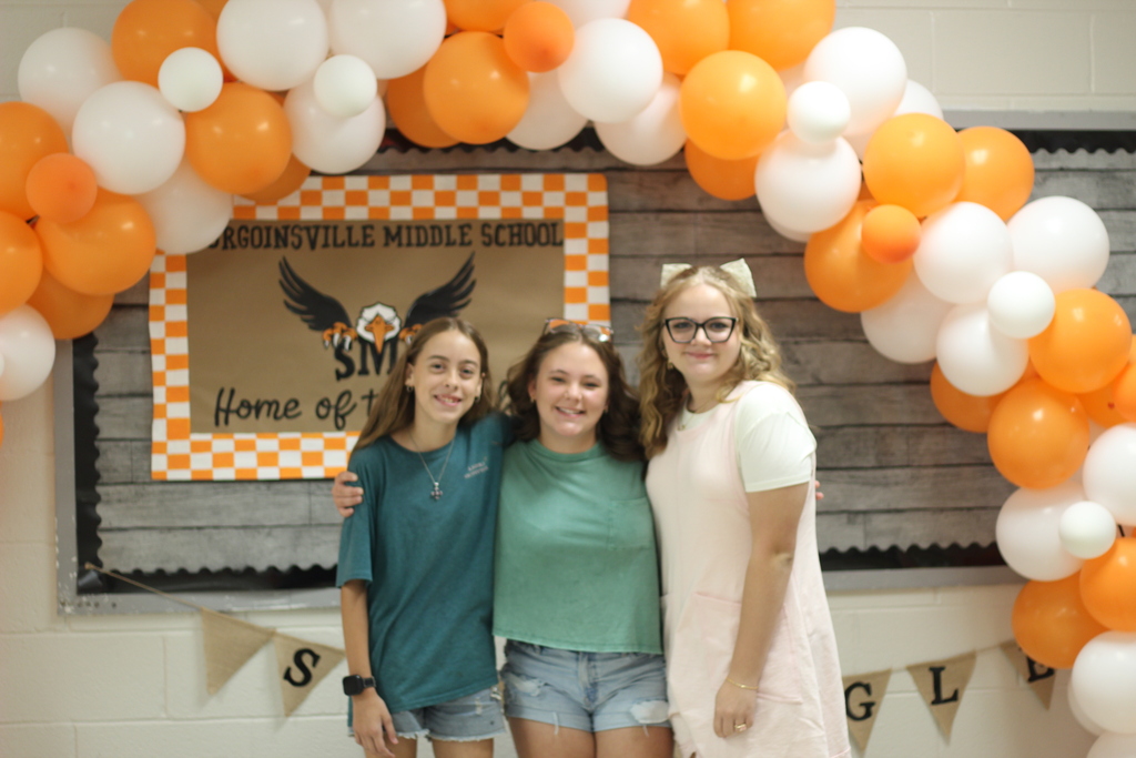 Three young women pose for a photo under a decorative balloon arch in front of a school banner.