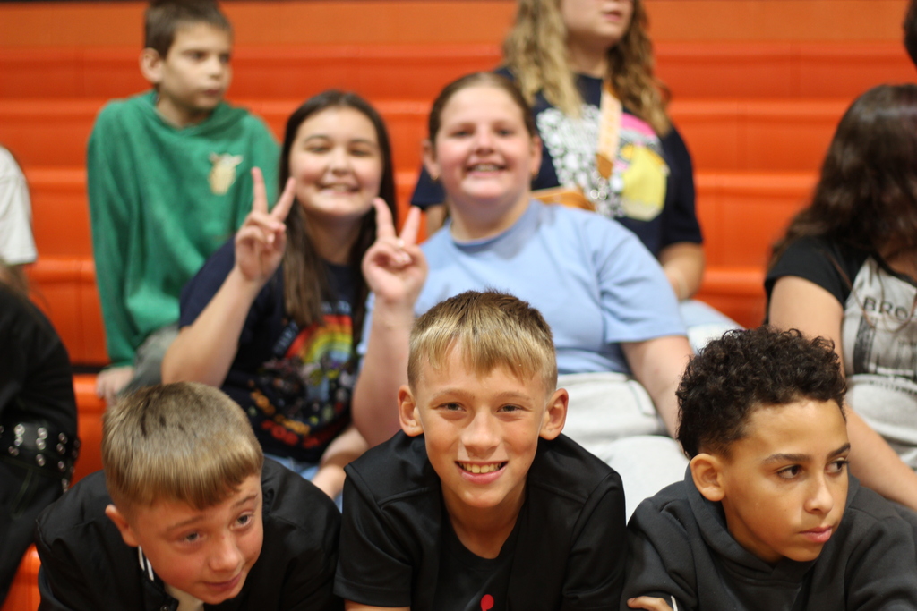 Several children seated on bleachers, some smiling and making peace signs, wearing various colored shirts.