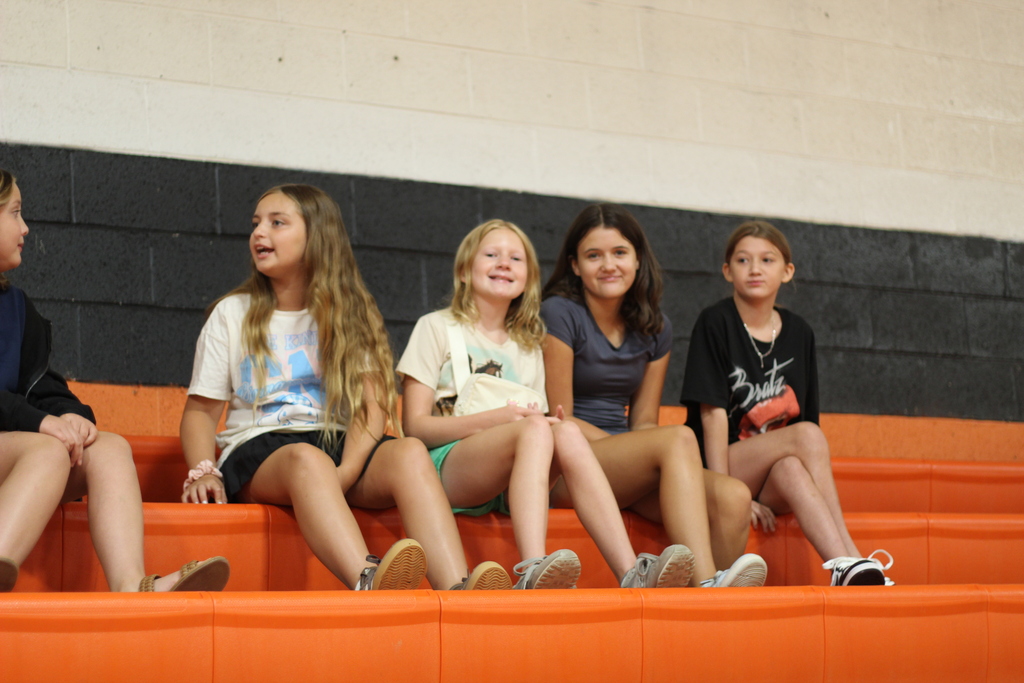 Five young girls sit on orange bleachers, smiling and looking forward, with a black and white wall in the background.
