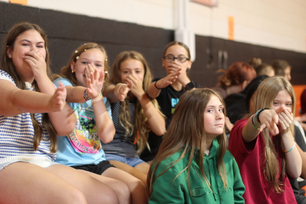 Several girls seated in a row, covering mouths with hands. One wears glasses. Orange wall in background.