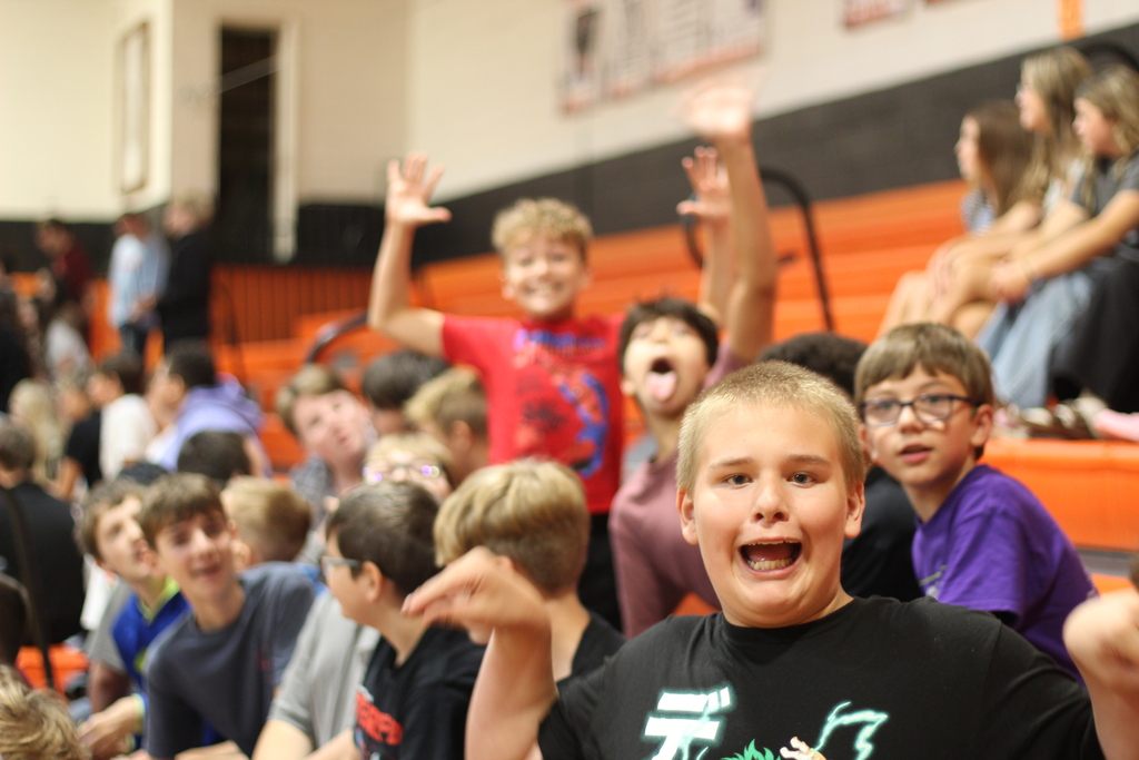 A crowd of children in a school gymnasium, some with hands raised. Two wearing glasses.