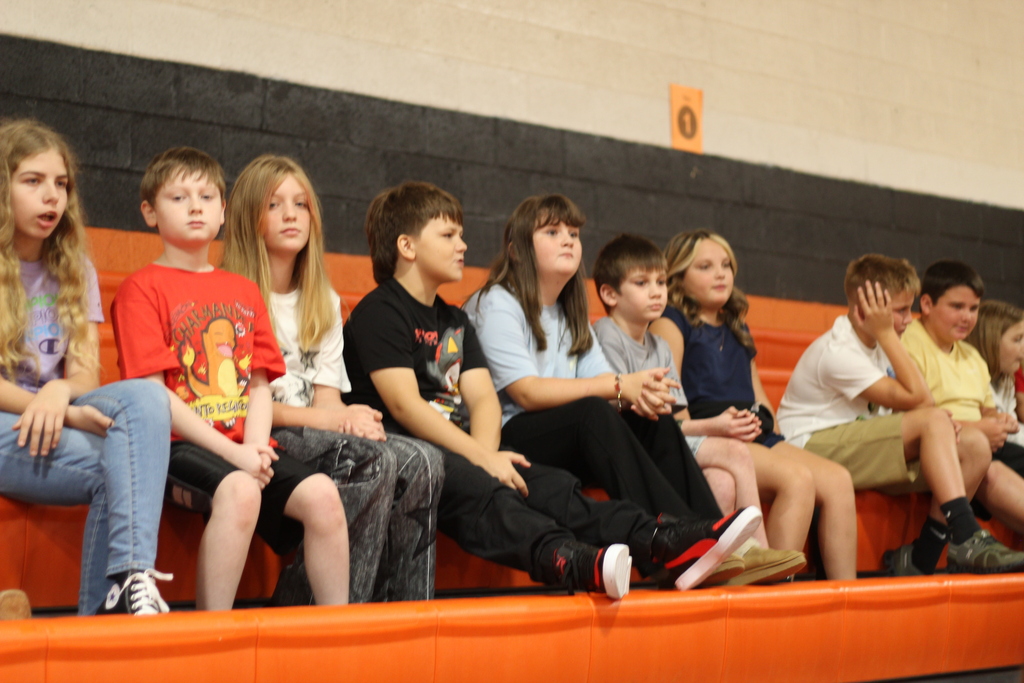 Several children in casual clothes sit on orange bleachers in a gym, facing forward, with one child in the middle wearing a red T-shirt.
