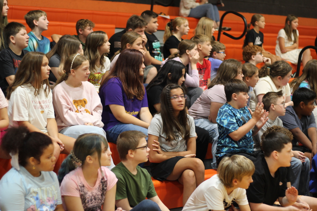 A crowd of children sitting in orange bleachers, some wearing glasses and others clapping, possibly during a school event.