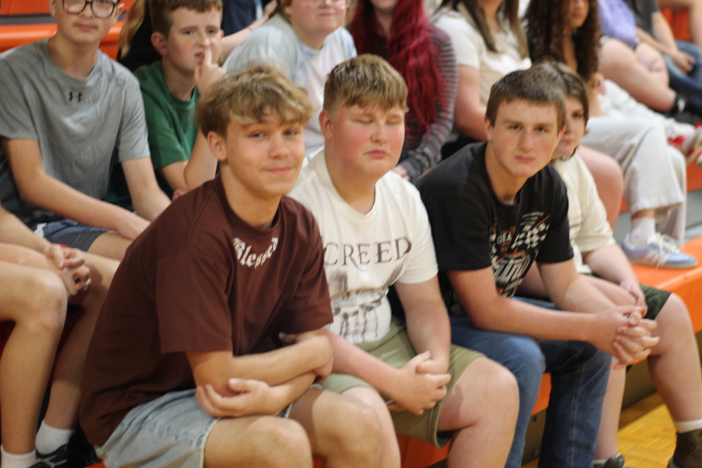 Four young men sit together on bleachers, each wearing casual clothing. One wears a brown shirt, one a white shirt, and the other two wear black.
