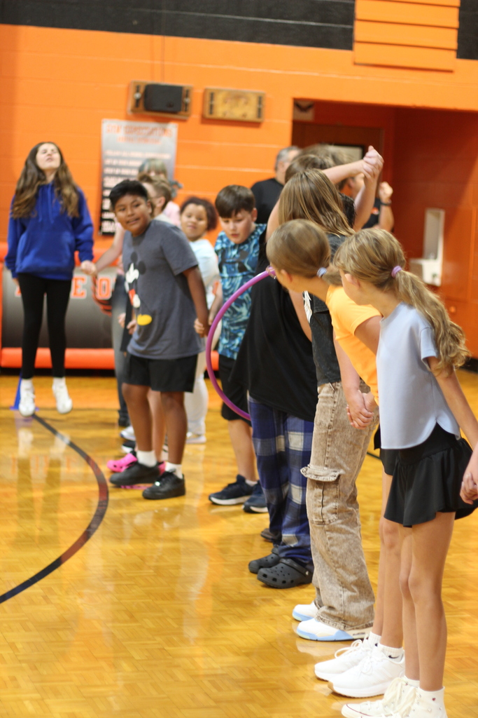 Several children and adults gather on a basketball court, some holding a hula hoop. An orange wall and black line mark the background.