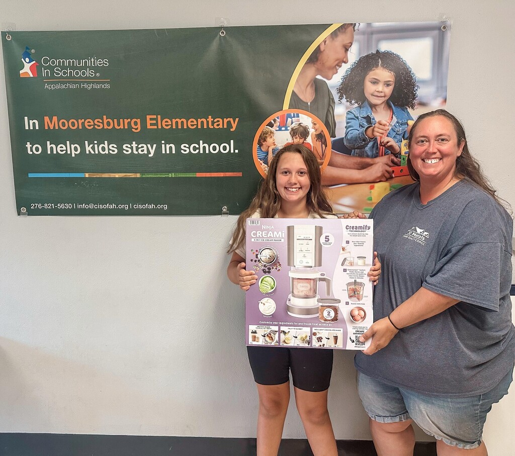 Two women stand in front of a banner with the text "Communities in Schools" and "In Mooresburg Elementary to help kids stay in school".