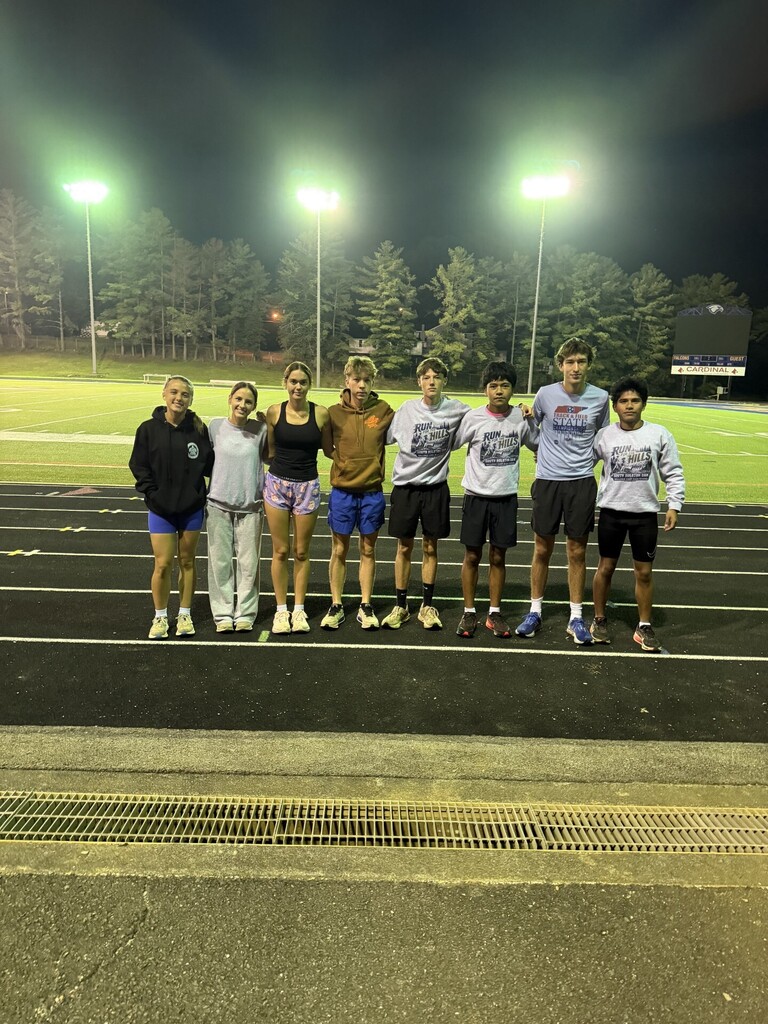 A group of eight athletes wearing sports gear, standing on a track with illuminated poles in the background.