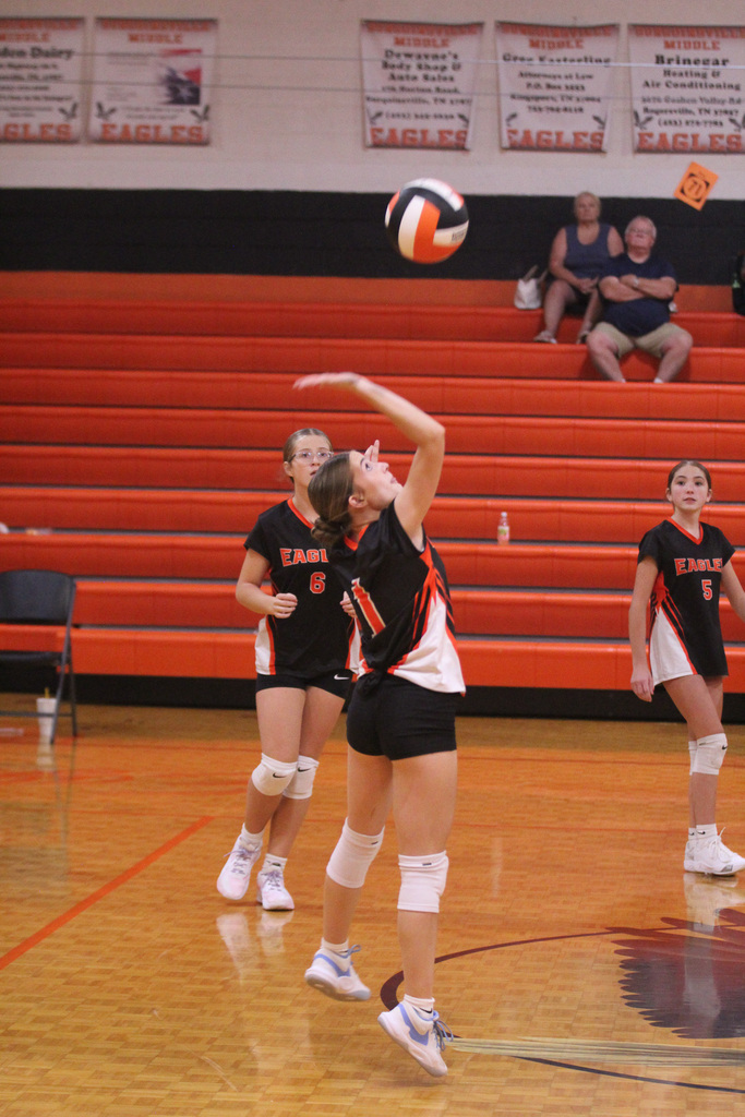 Three women in matching uniforms play volleyball. One player jumps to hit the ball. Behind, spectators sit on red bleachers.