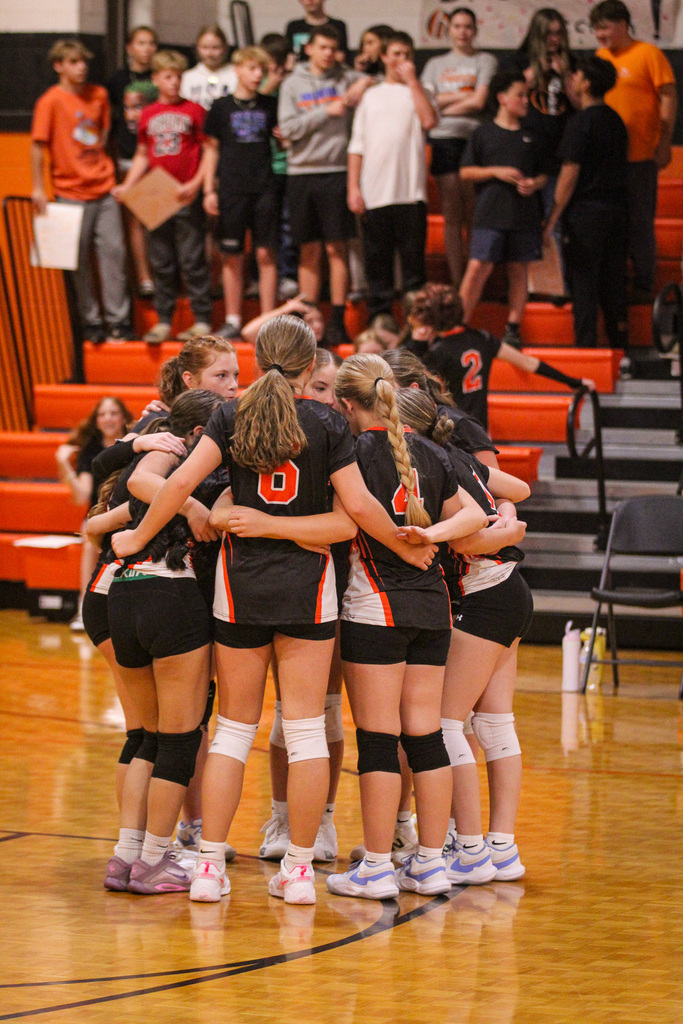 A group of female volleyball players with numbers on their jerseys, standing together in a huddle on a basketball court.