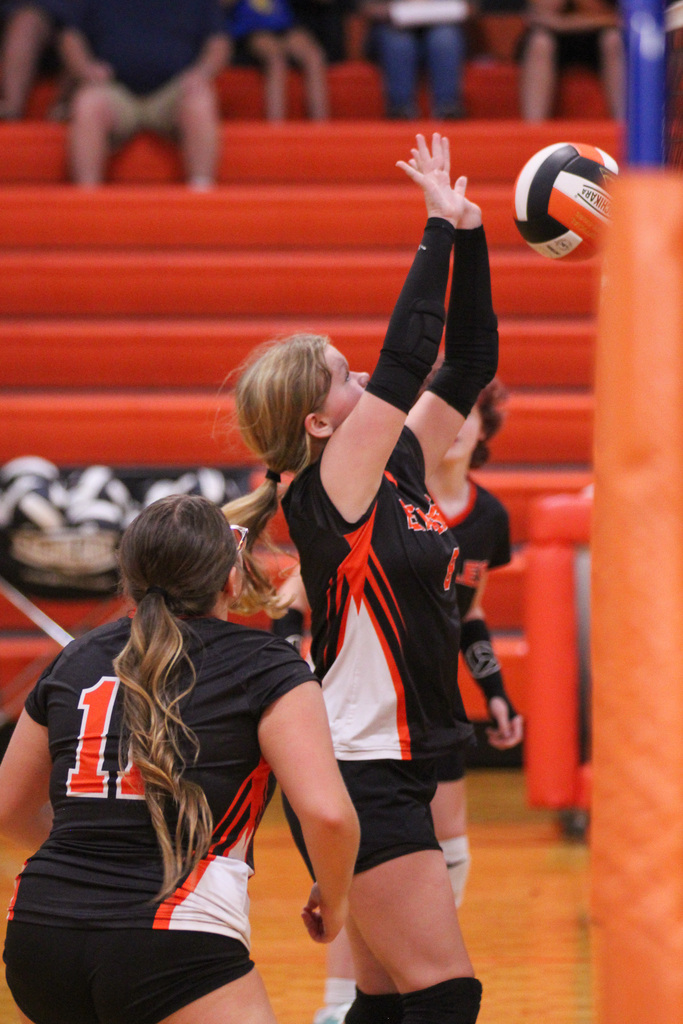 Volleyball players in black uniforms with numbers, one jumping to hit the ball. Spectators in red bleachers.