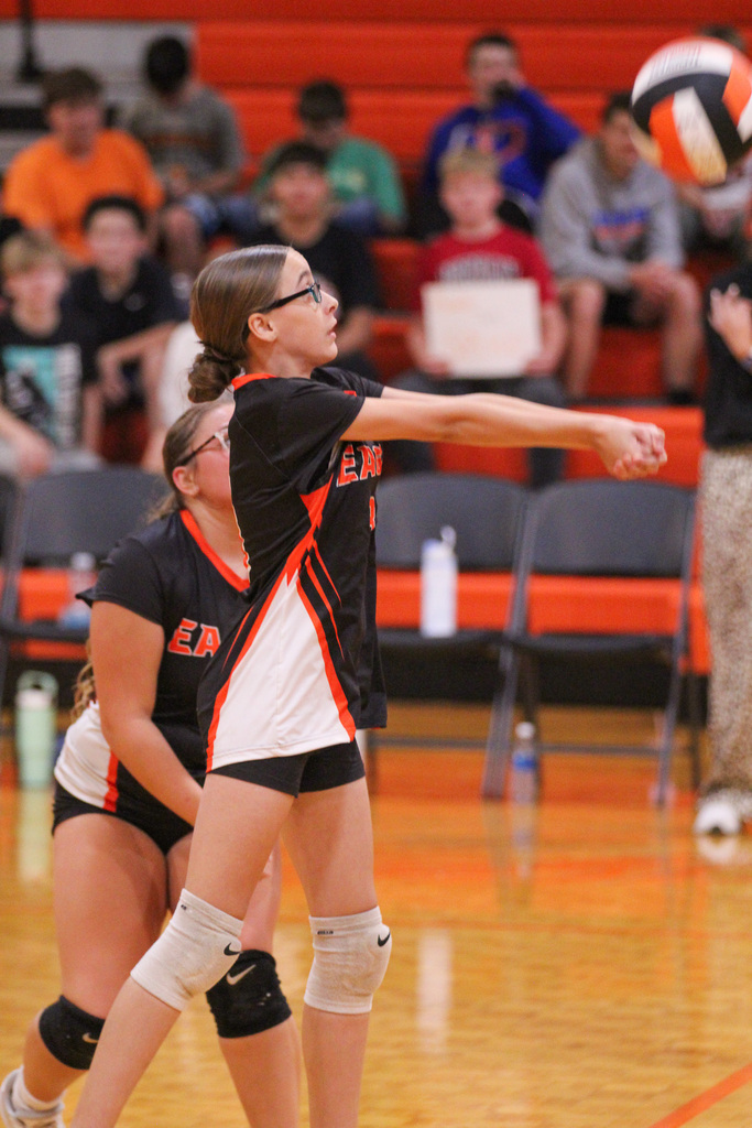 Two women play volleyball on a court. One woman in glasses holds a ball. Behind, spectators watch from red bleachers.