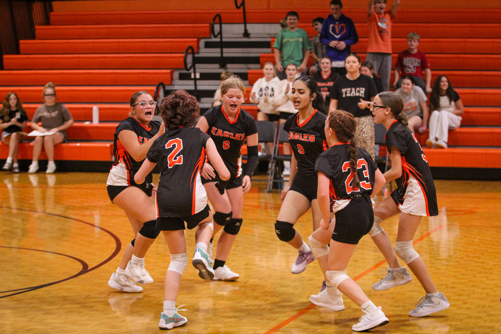 Team in black uniforms celebrates on a basketball court. Background shows bleachers with seated spectators.
