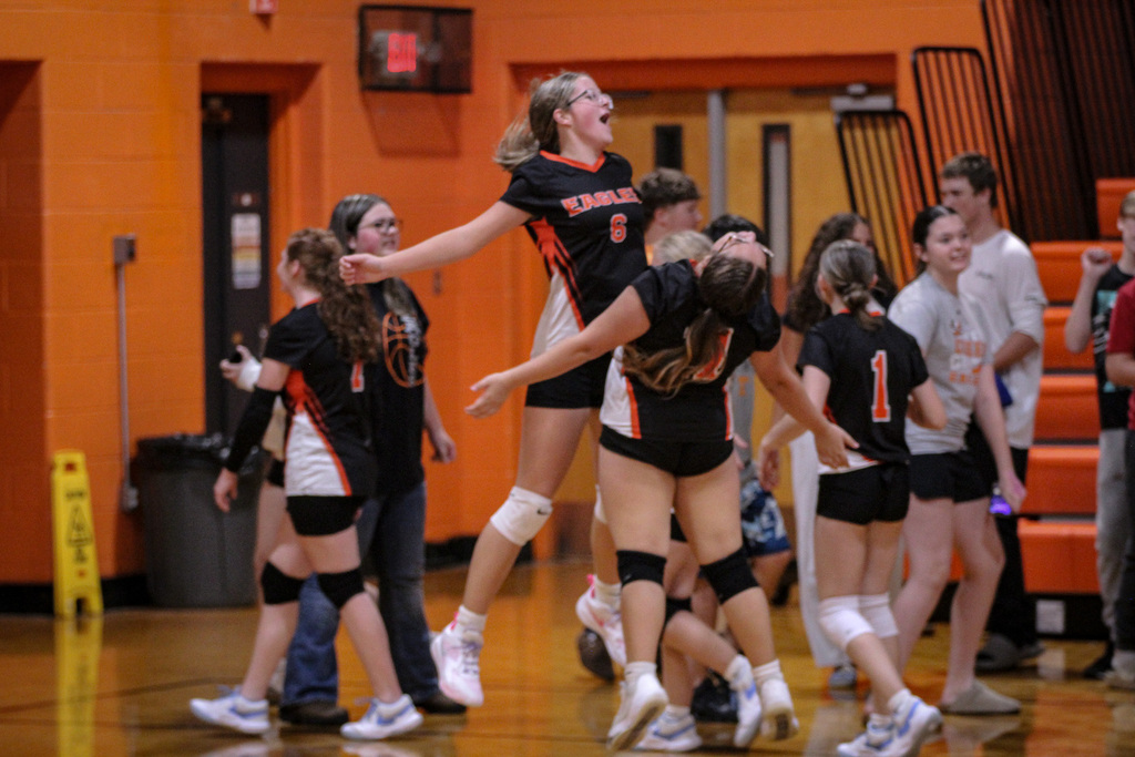 Indoor volleyball players celebrate a successful play. Some players jump while others cheer in a gym with orange walls.