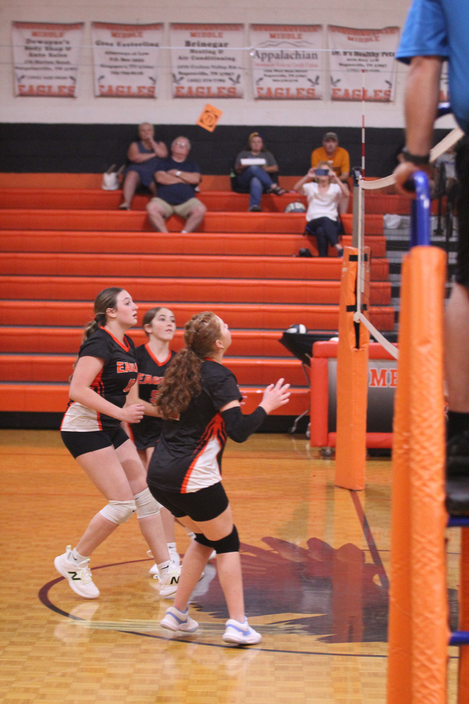 Four players in black uniforms on a court with a volleyball net. Spectators in the stands.