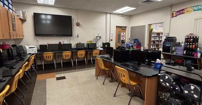 An empty room with computers, chairs, a flat-screen monitor on a wall, and white carpet.